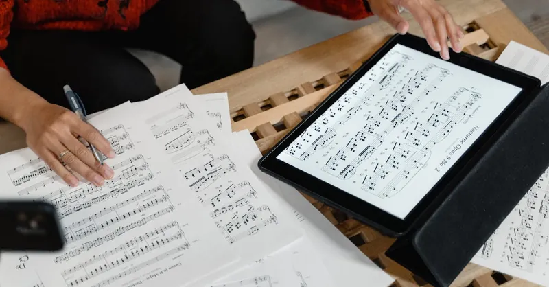 A person reviewing and composing sheet music using both paper and digital formats on a table.