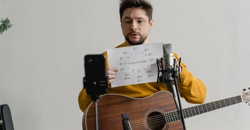 Man with acoustic guitar explains music theory online using smartphone and microphone.