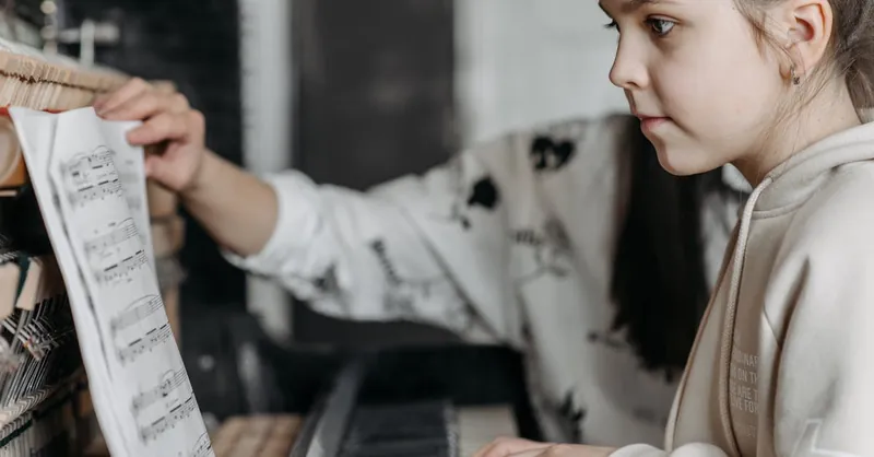 Girl in hoodie learning to play piano with sheet music during indoor lesson.