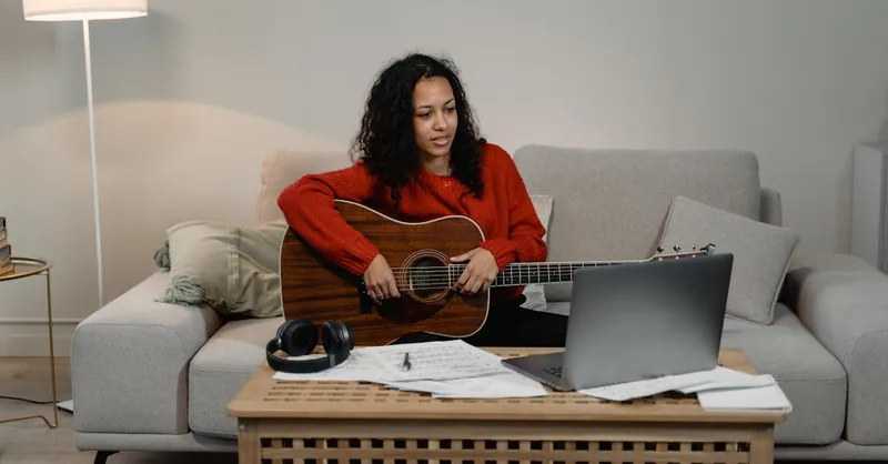 Woman in living room playing guitar while learning online via laptop. Cozy and creative setup.