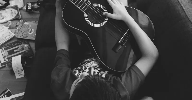 A young boy lying indoors on a couch playing an acoustic guitar with scattered books around.