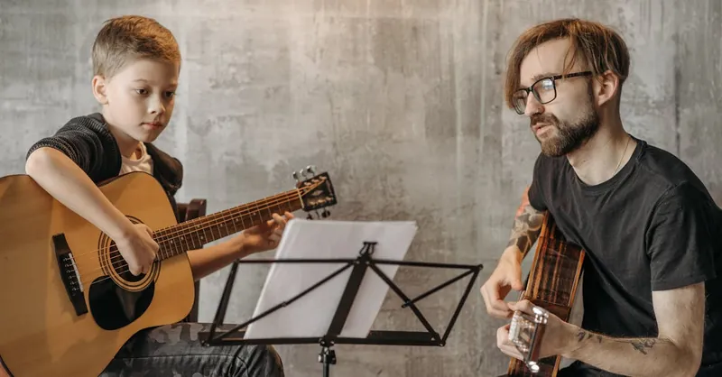 A young boy learns guitar from a teacher during a lesson indoors.