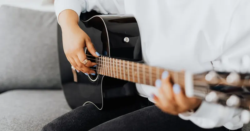 Person playing an acoustic guitar indoors, focusing on the fretboard and hands.