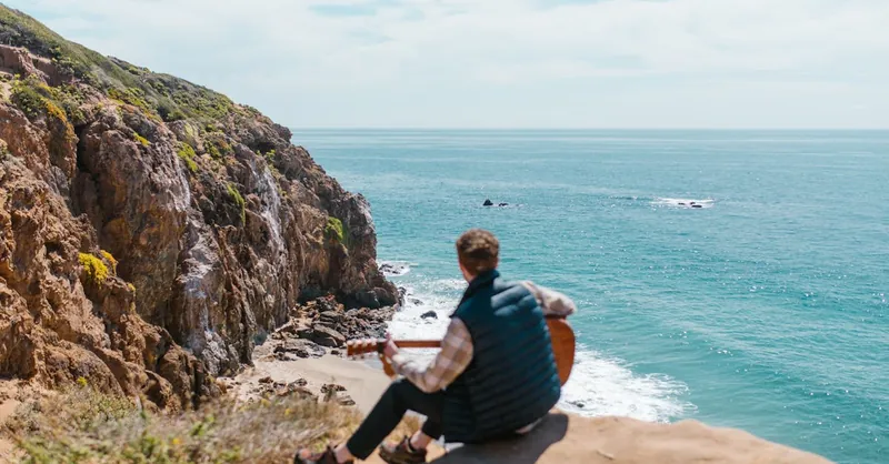 Back view of a man playing guitar on a cliff by the ocean under clear skies.