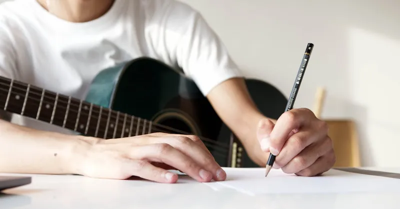 Guitarist writing song notes while holding acoustic guitar, emphasizing creativity and practice.